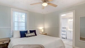 Ein Schlafzimmer mit Bett, Deckenventilator, Nachttisch, Fenster und Blick ins Badezimmer.