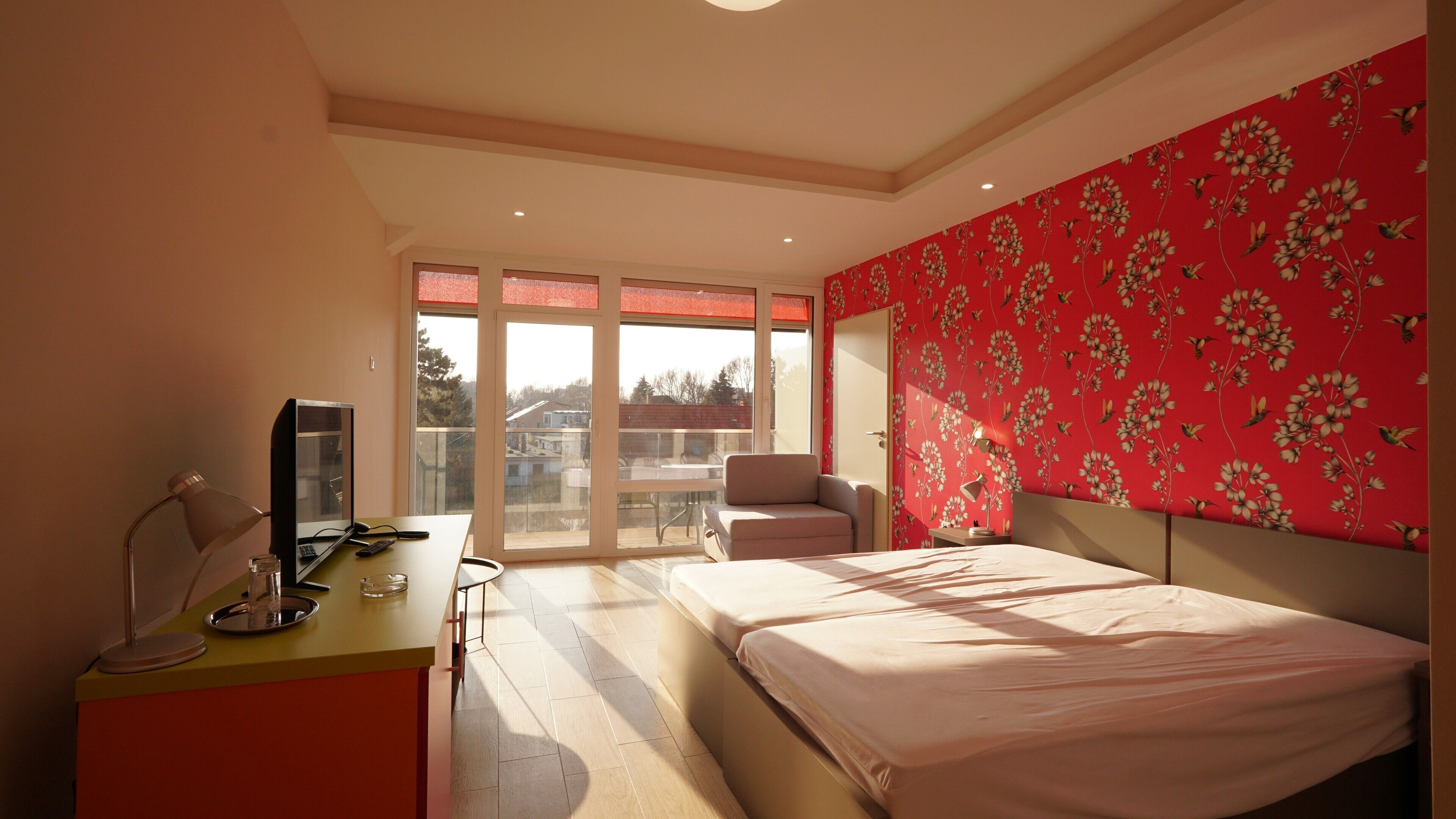 Ein Schlafzimmer mit einem Bett, einem Schreibtisch mit Computer, einem Stuhl und einem Fenster mit Blick auf Gebäude.