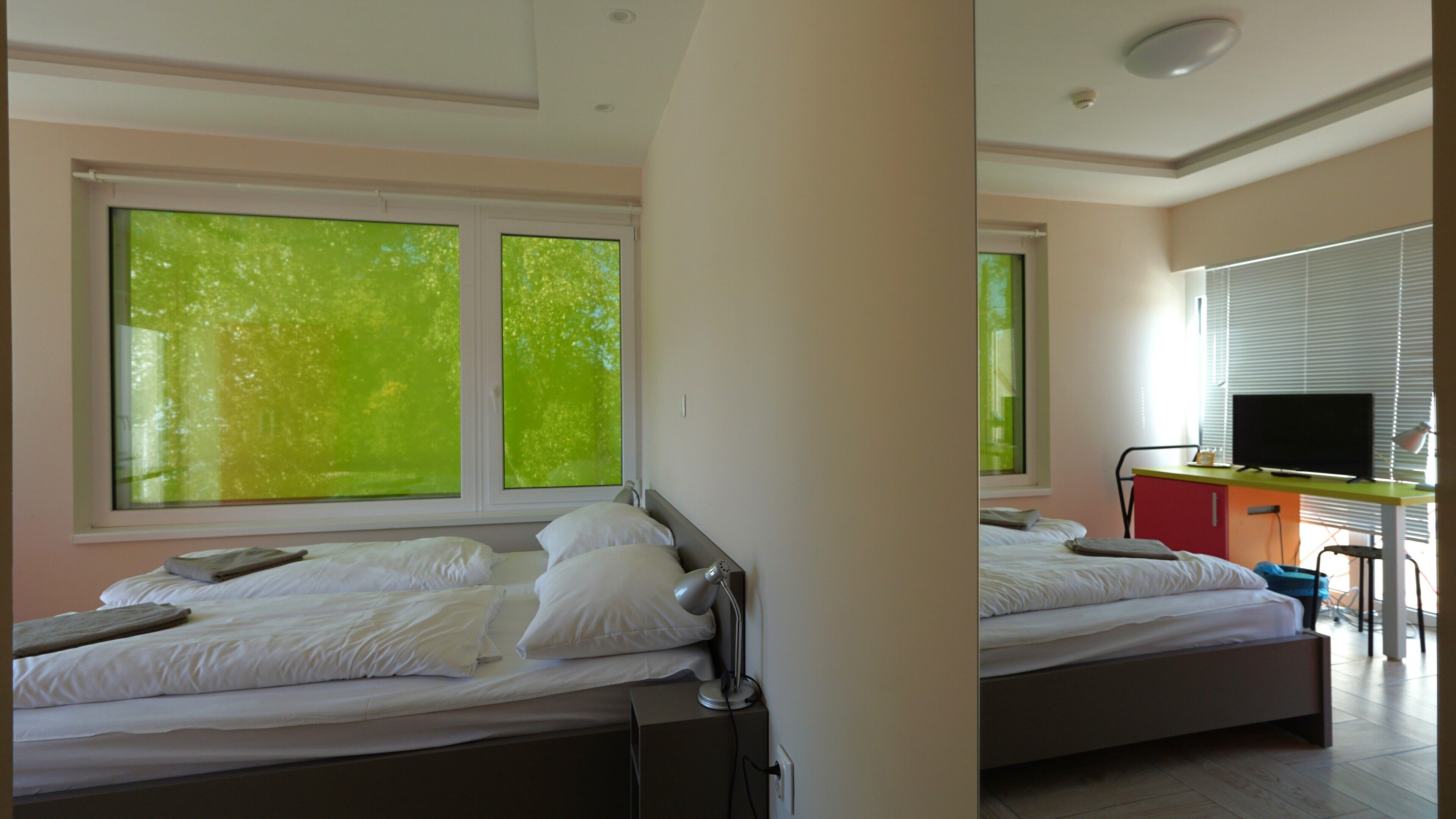 A bedroom with two beds, a desk with a TV, and large windows with a view of trees.