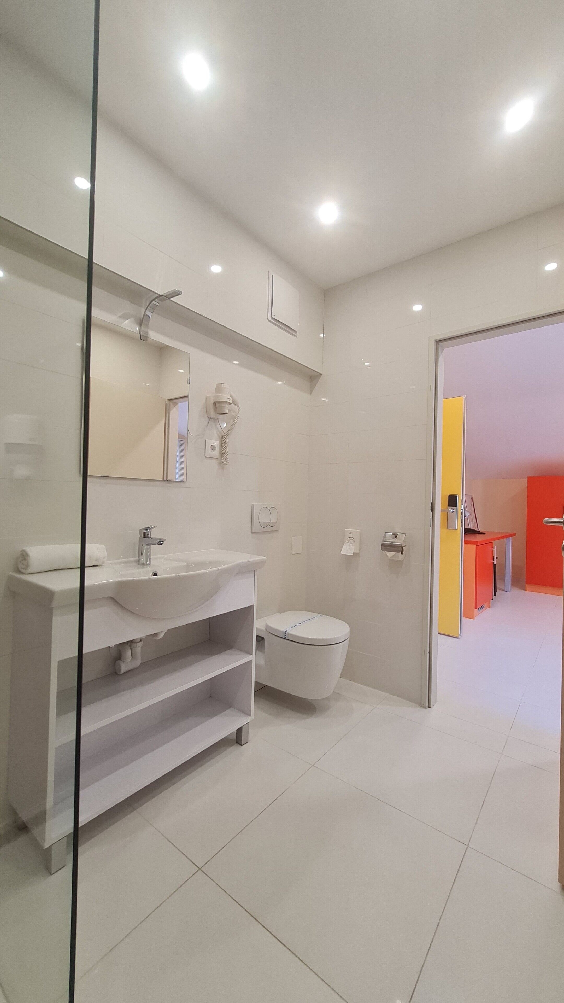 A modern bathroom with a white sink, mirror, and toilet. There is a glass partition on the left, and a red door in the background.