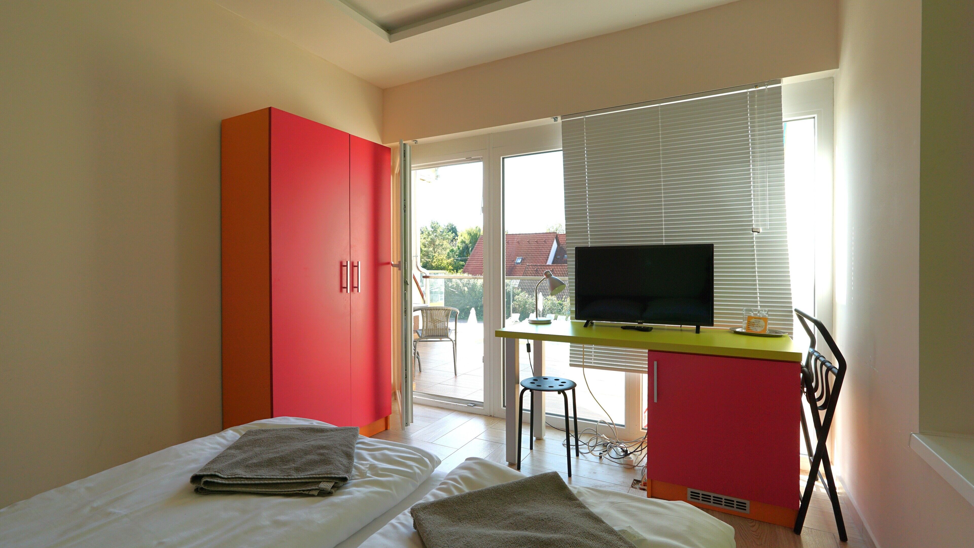 A bedroom with a bed, a desk with a TV, a chair, and a window with blinds.