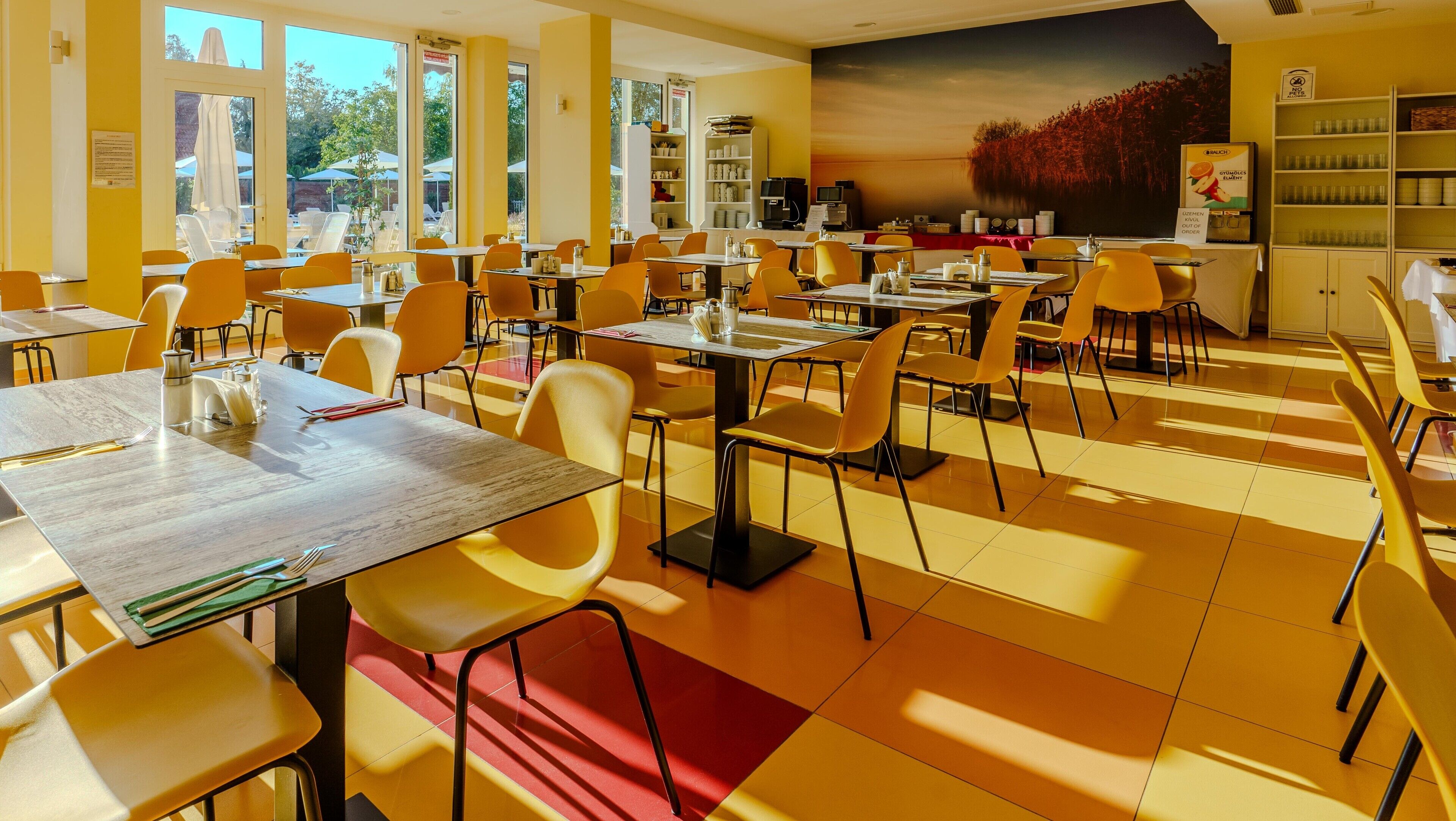 A dining area with yellow chairs and tables, large windows, and a mural on the wall.