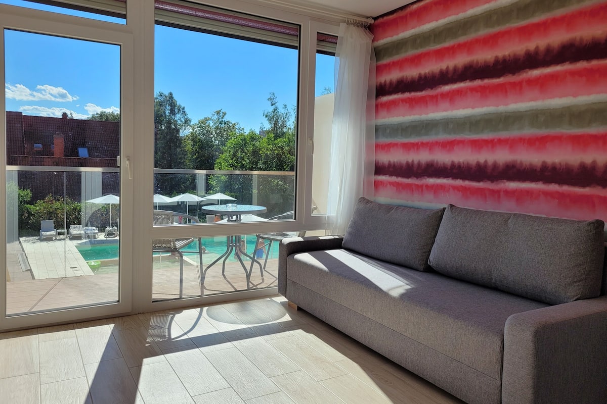 A modern living room with a sofa, a view of a pool, and a balcony with outdoor furniture.