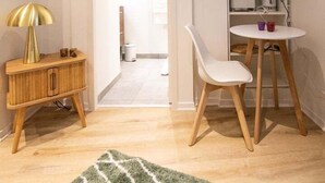 A compact living space with a wooden side table, a green patterned rug, a white chair, a small round table with cups, and a TV mounted on the wall. - Nice little studio near Opera (Lyon)