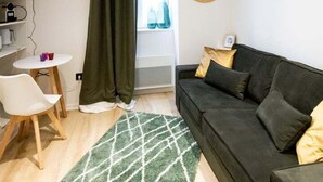 A compact living space with a dark sofa, a small round table, a TV mounted on the wall, and a window with curtains. - Nice little studio near Opera (Lyon)