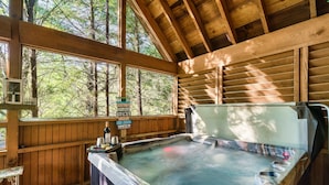 Eine Holzhütte mit Whirlpool, einer Holzterrasse und großen Fenstern mit Blick auf einen Wald.