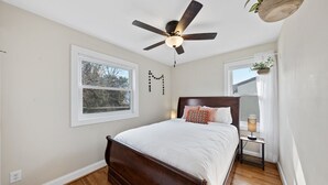 A bedroom with a wooden bed, a ceiling fan, a bedside table with a lamp, and a window with a view of trees. - Charming 3-Bed Bungalow with Fenced-in Yard (Raleigh)