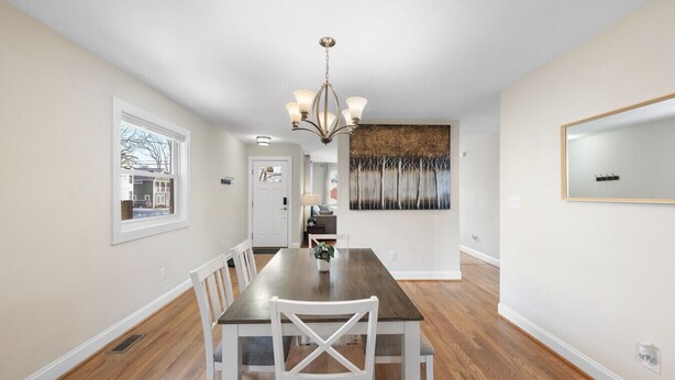 A dining area with a long wooden table, white chairs, a chandelier, and a large abstract painting on the wall. - Charming 3-Bed Bungalow with Fenced-in Yard (Raleigh)