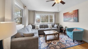 A living room with a sectional sofa, a coffee table, a blue armchair, a patterned rug, and a ceiling fan. - Charming 3-Bed Bungalow with Fenced-in Yard (Raleigh)