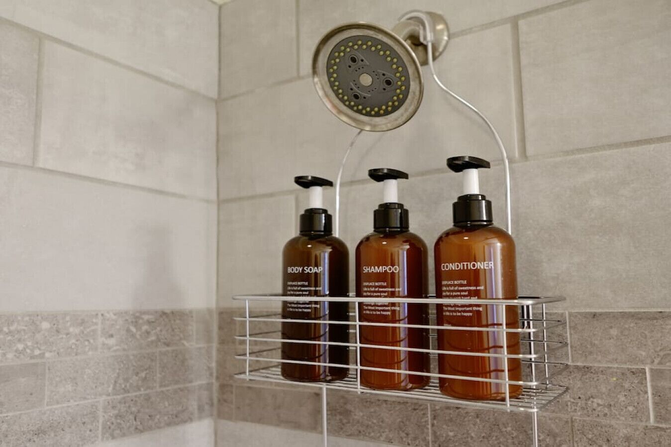 A bathroom with a shower, a wall-mounted shelf holding three bottles of body products, and a showerhead.