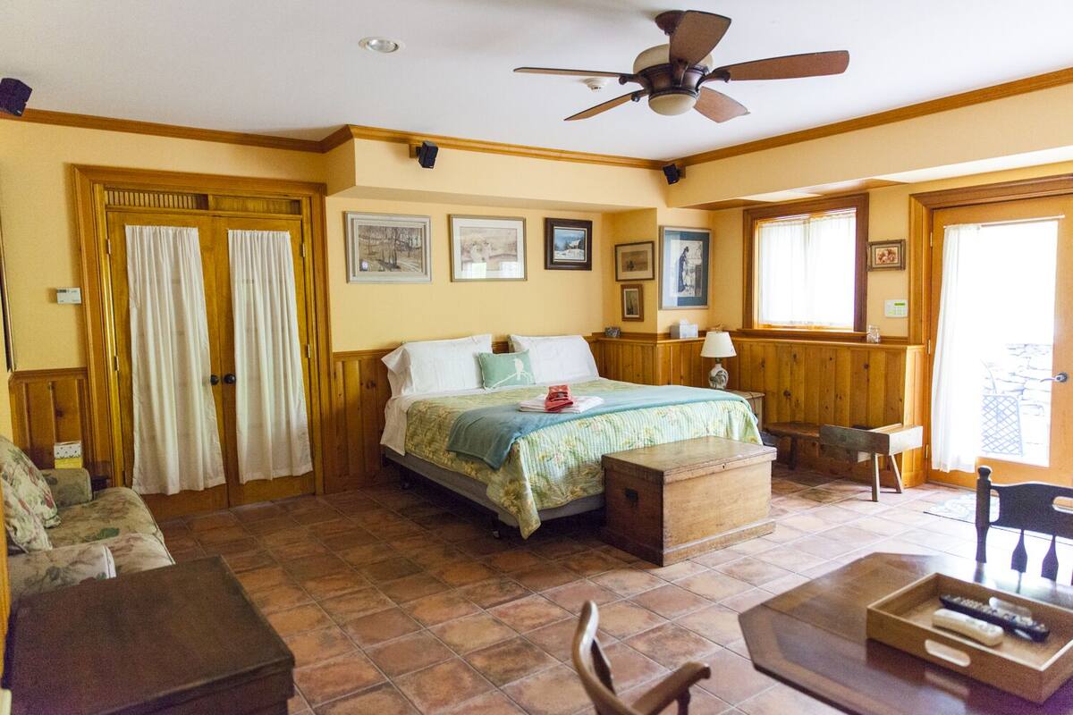 Um quarto aconchegante com uma cama, um banco de madeira, uma mesa com controles remotos e um ventilador de teto.