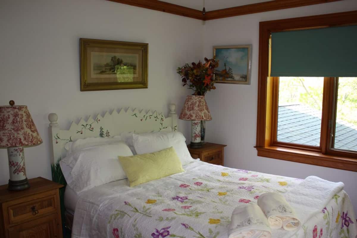 Quarto com colcha floral, cabeceira branca, travesseiro amarelo e janela com vista para o telhado.