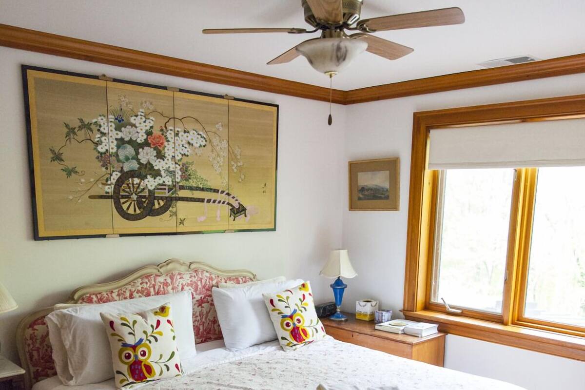 Quarto com cama, ventoinha no teto, janela e um quadro na parede.