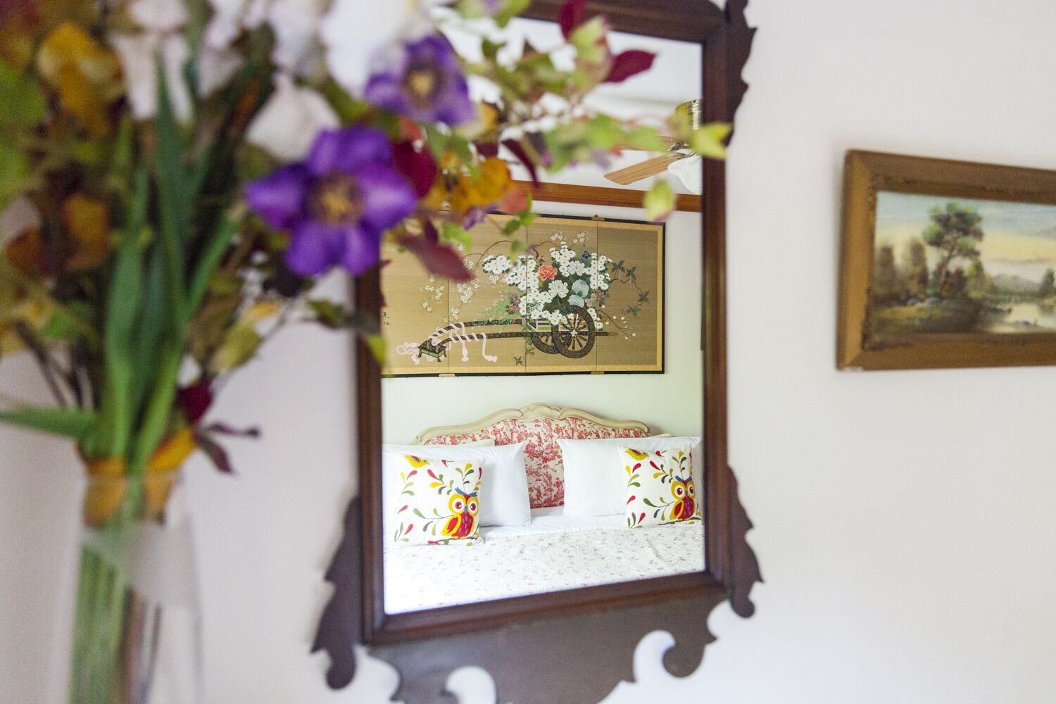 A mirror reflecting a bed with floral bedding and an owl-themed pillow, a framed painting, and a vase with flowers.