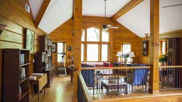 A spacious cabin with a high ceiling, wooden beams, a library, a dining area, and large windows.