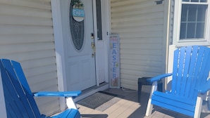 Eine Veranda mit zwei blauen Adirondack-Stühlen, einer weißen Tür mit Glasfenster und einem Schild mit der Aufschrift 'Happy Easter'.