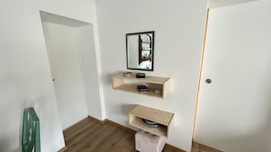 A room with wooden flooring, a green chair, and a white door. There are two wooden shelves with various items on them. - Idyllischer Bauernhof 3 (Wildensbuch)
