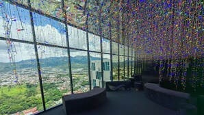 A modern interior with large windows offering a panoramic city view, a seating area with a curved bench, and a high ceiling adorned with hanging lights.