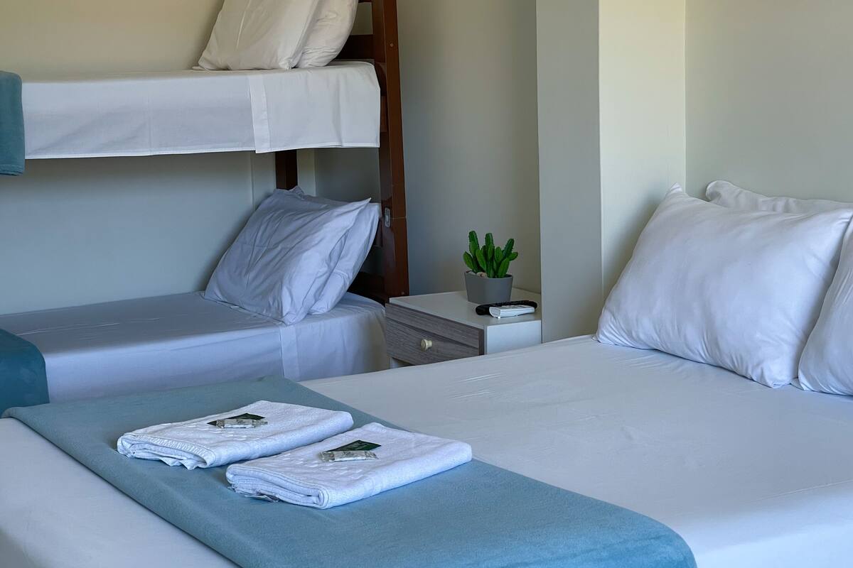 A hotel room with a bunk bed, two single beds, a bedside table with a remote control, a potted plant, and folded towels on the beds.