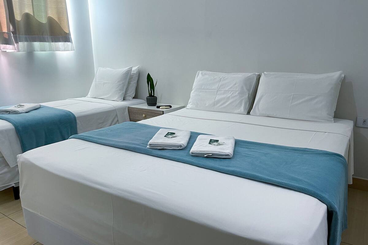 A hotel room with two single beds, a large bed, a nightstand with a lamp, and a potted plant.