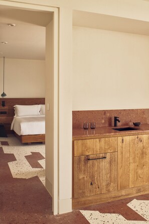A modern hotel room with a wooden bed, a kitchenette with a sink and cabinets, and a patterned floor. - Nous Santorini (Santorini)