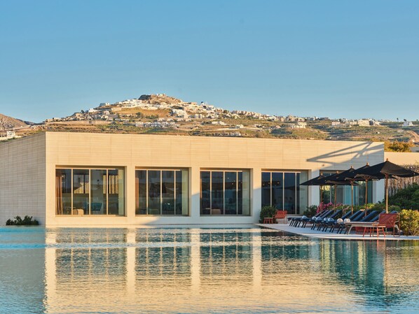 A modern building with a pool, large windows, and a view of a hill with buildings. - Nous Santorini (Santorini)