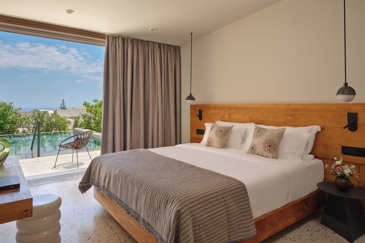 A bedroom with a large bed, a view of the ocean, and a balcony with a chair.