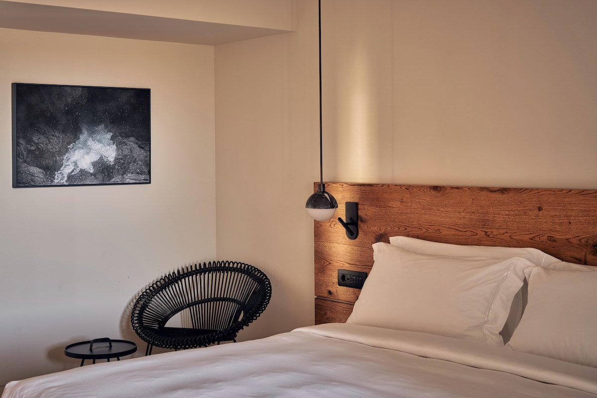 A bedroom with a bed, a chair, a small table, a wall-mounted light, and a framed picture.