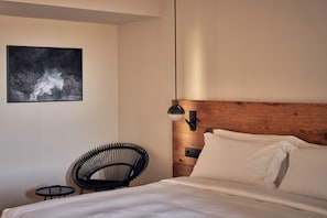 A bedroom with a bed, a chair, a small table, a wall-mounted light, and a framed picture. - Nous Santorini (Santorini)