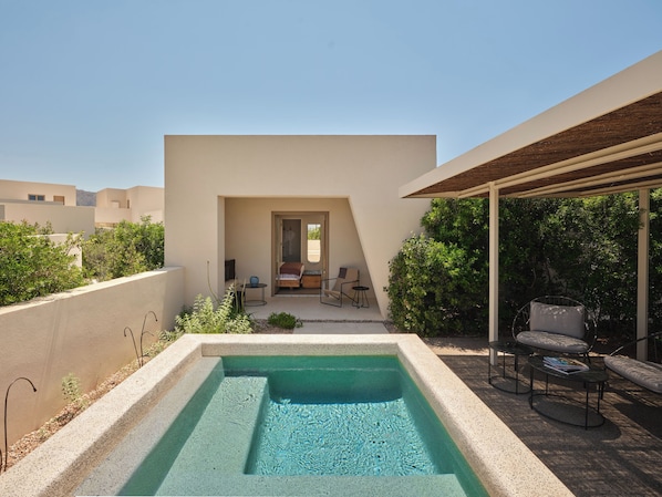 A modern outdoor pool area with a clear view of the pool, a covered seating area, and a pathway leading to a building entrance. - Nous Santorini (Santorini)