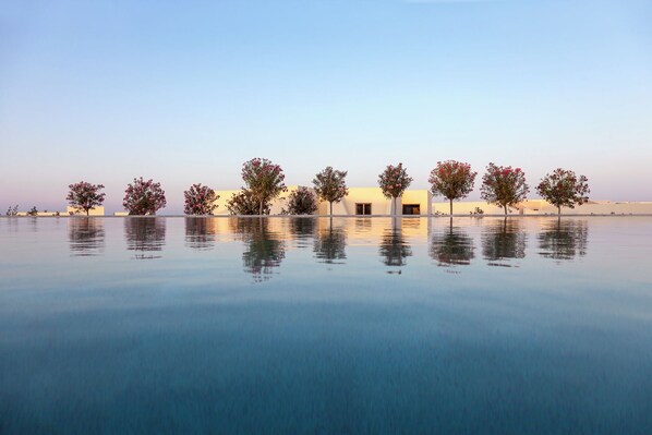 A serene pool area with a row of trees and a building in the background. - Nous Santorini (Santorini)