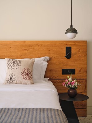 A bedroom with a wooden headboard, a bed with white bedding, a pillow with floral embroidery, a bedside table with a vase of flowers, and a hanging light fixture. - Nous Santorini (Santorini)