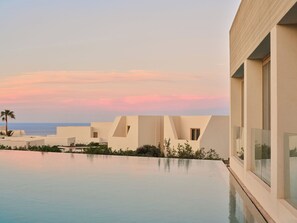 A modern resort with a pool, white buildings, and a view of the ocean at sunset. - Nous Santorini (Santorini)