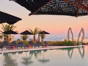 A poolside area with lounge chairs and umbrellas, palm trees, and a view of the ocean. - Nous Santorini (Santorini)