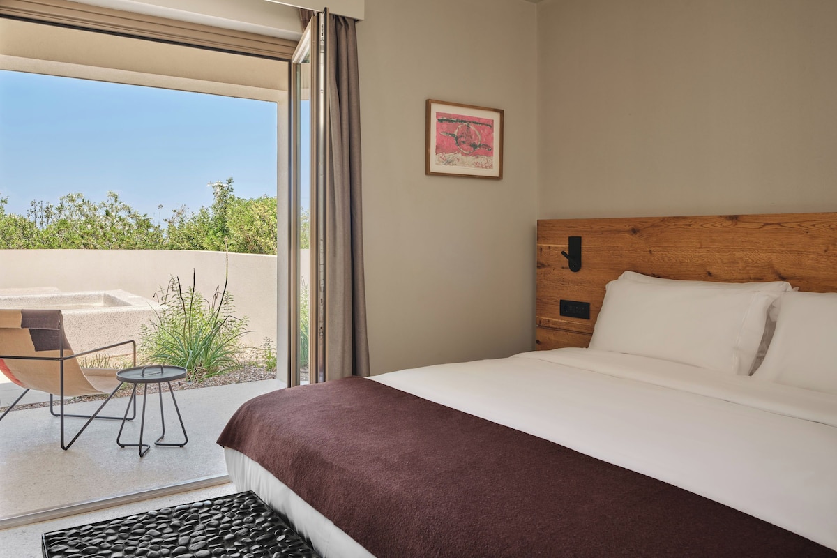 A hotel room with a bed, a small table, a chair, a window with curtains, and a picture on the wall.