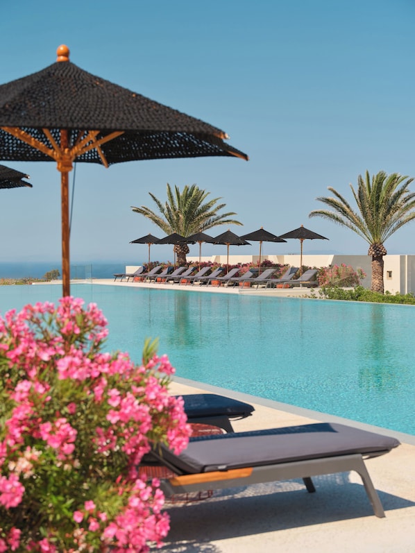 A poolside area with lounge chairs and umbrellas, a clear sky, and a view of the sea. - Nous Santorini (Santorini)