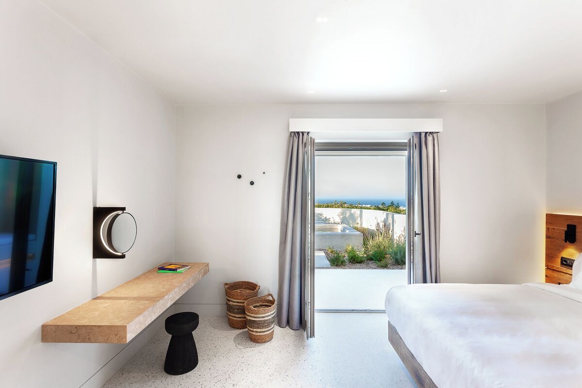 A modern hotel room with a bed, a TV, a bedside table, a mirror, and a view of the outdoors.