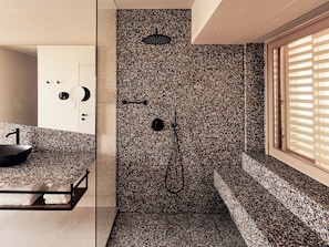 A modern bathroom with a granite countertop, a black sink, a shower with a black handle, and a window with blinds. - Nous Santorini (Santorini)