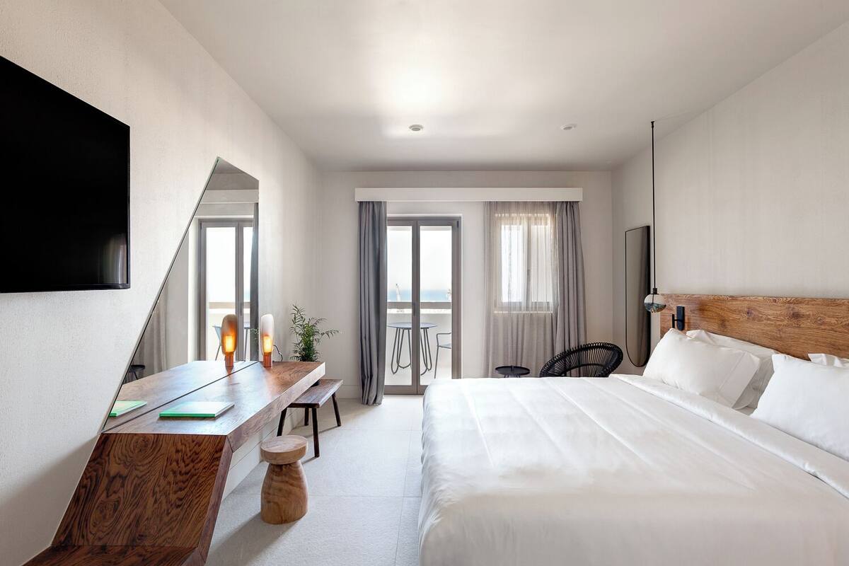 Habitación de hotel con cama, escritorio, silla, televisor y balcón con vistas.