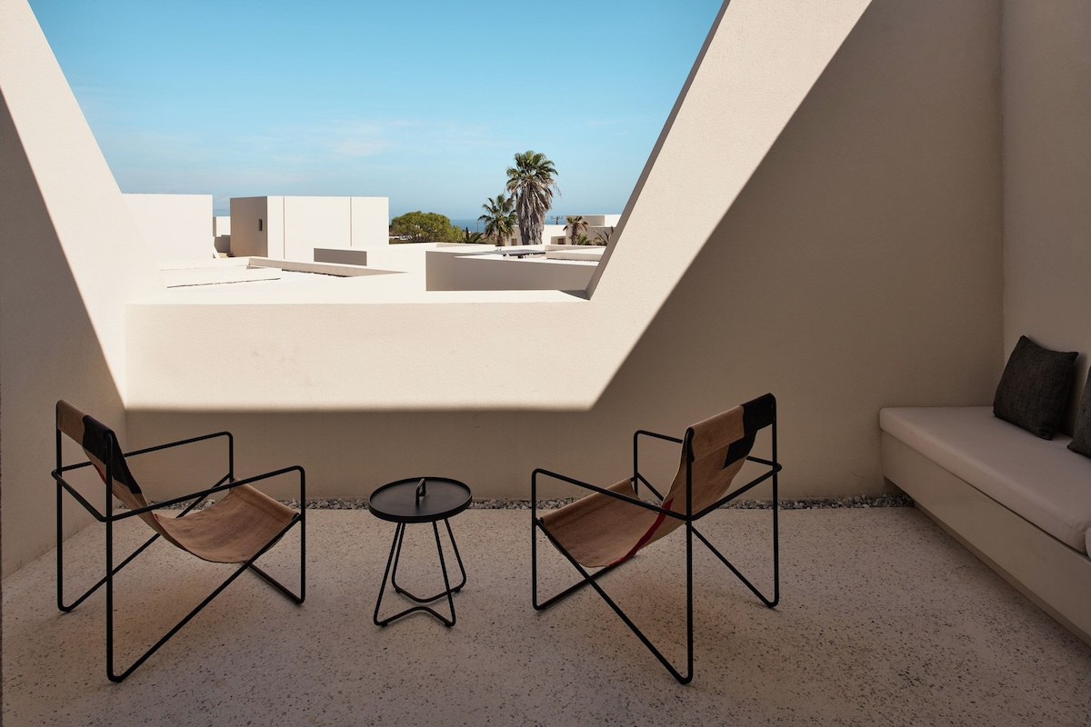 A modern outdoor seating area with a table and two chairs, a sofa, and a view of the sea through a triangular skylight.