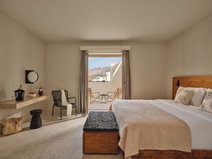 A bedroom with a bed, a wooden headboard, a bedside table, a chair, a small table, a mirror, and a view of the outdoors. - Nous Santorini (Santorini)