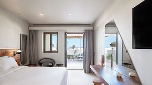 A hotel room with a large bed, a wooden nightstand, a mirror, and a view of the outdoor terrace. - Nous Santorini (Santorini)