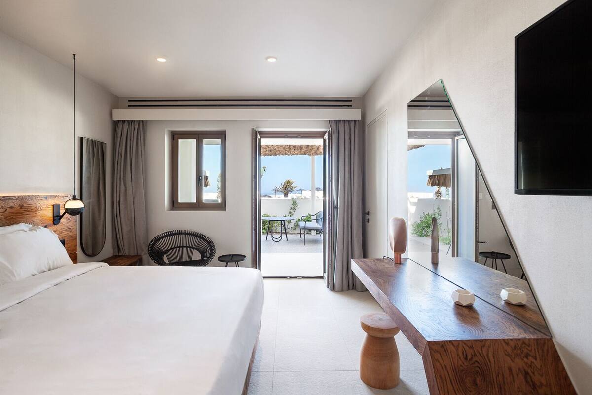 Habitación de hotel con una cama grande, una mesita de noche de madera, un espejo y vistas a la terraza exterior.