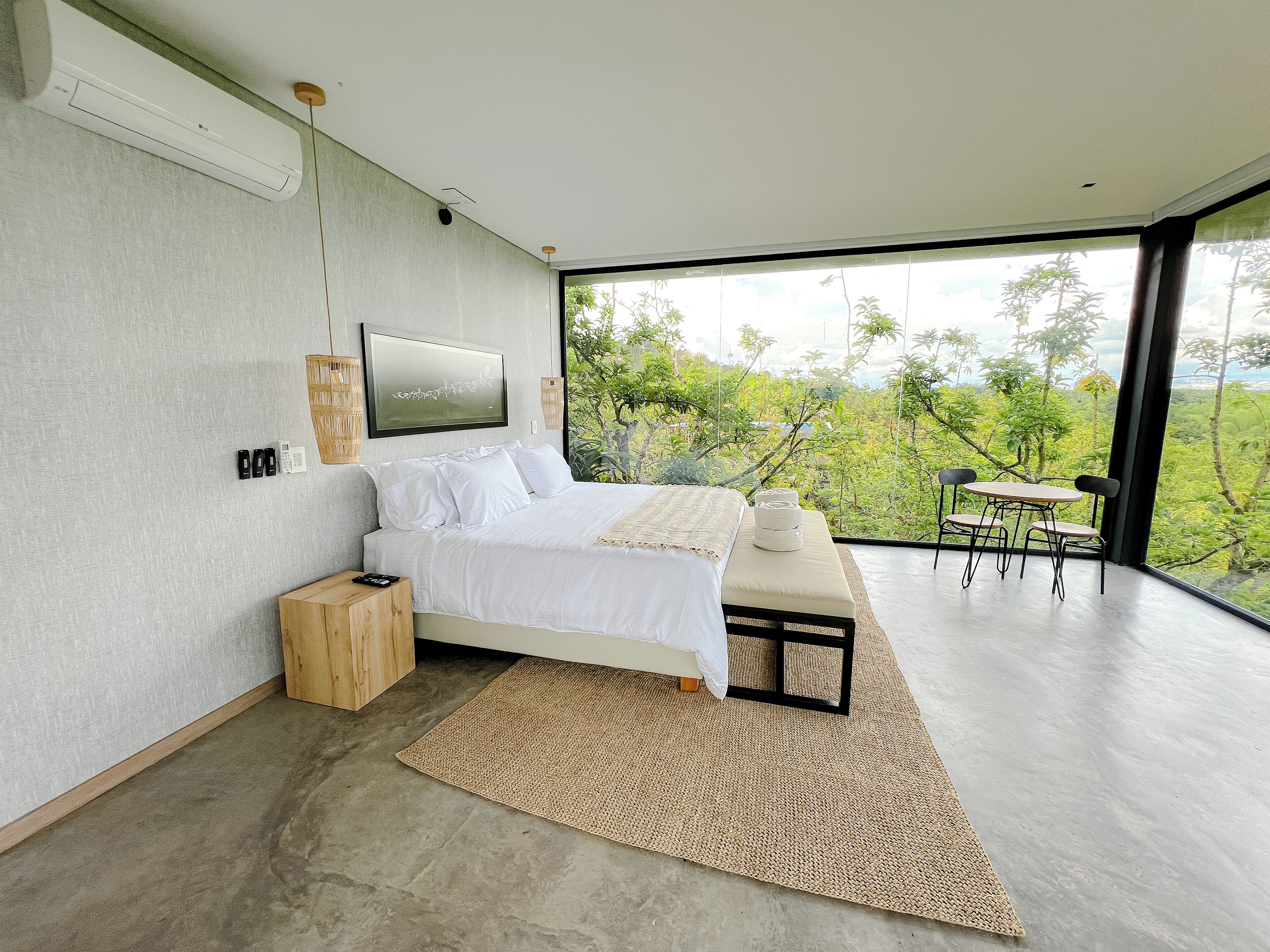 A modern bedroom with a large bed, a small table, and a view of lush greenery.
