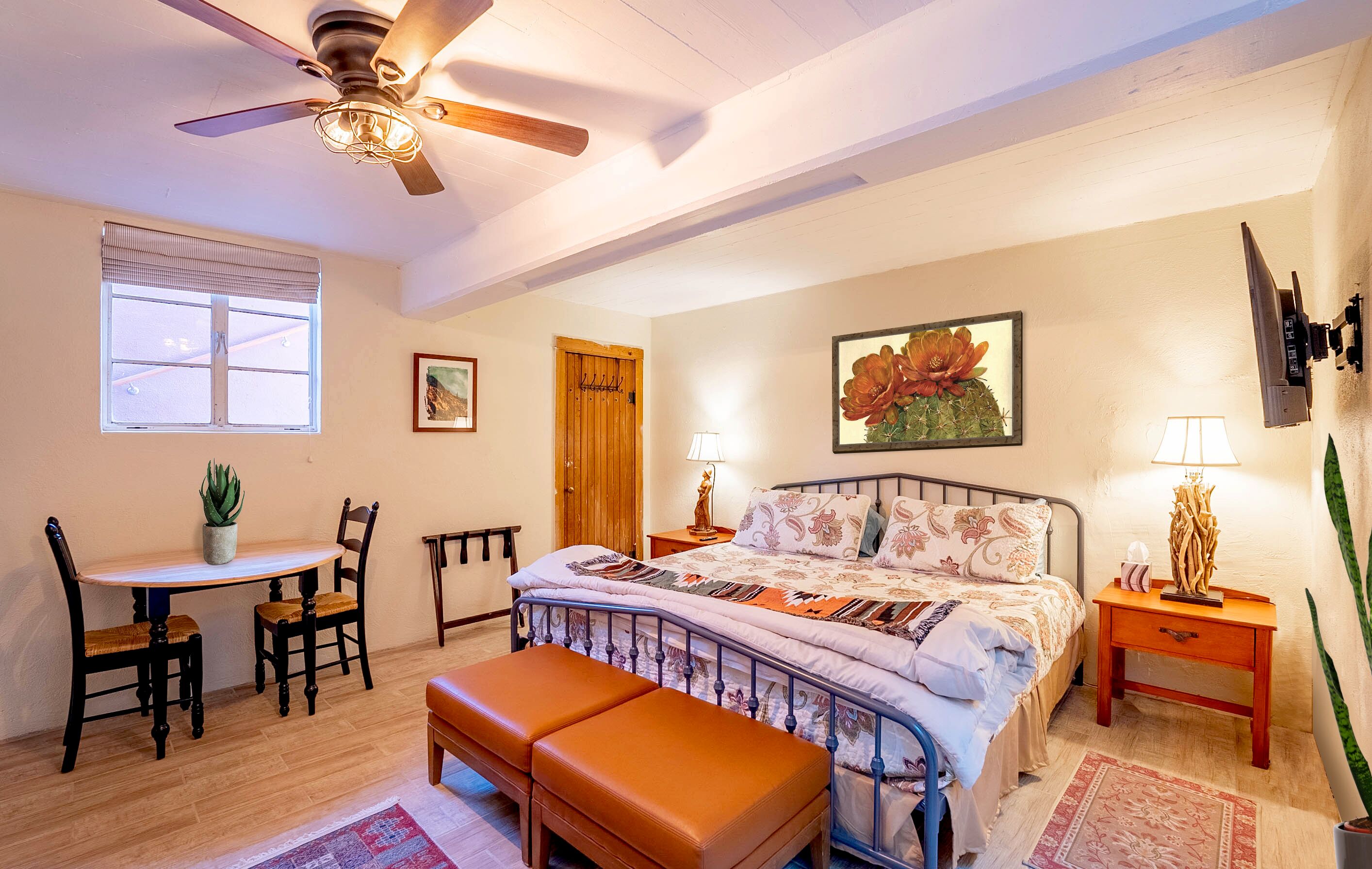 A bedroom with a bed, bedside tables, a desk with chairs, a ceiling fan, a window, and a door.