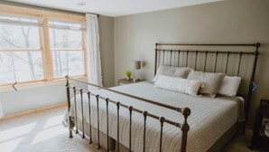 A bedroom with a metal bed frame, a large window with curtains, a bedside table with a lamp, and a nightstand. - Cozy Vacation Cottage on the Lake with Hot Tub!  (Webster)