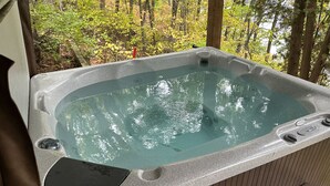 A hot tub with a stone exterior, surrounded by a wooden deck and a forested area. - Cozy Vacation Cottage on the Lake with Hot Tub!  (Webster)