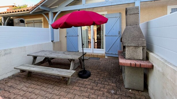 Un espace patio avec une table et un banc en bois, un parasol rouge et un barbecue en pierre.