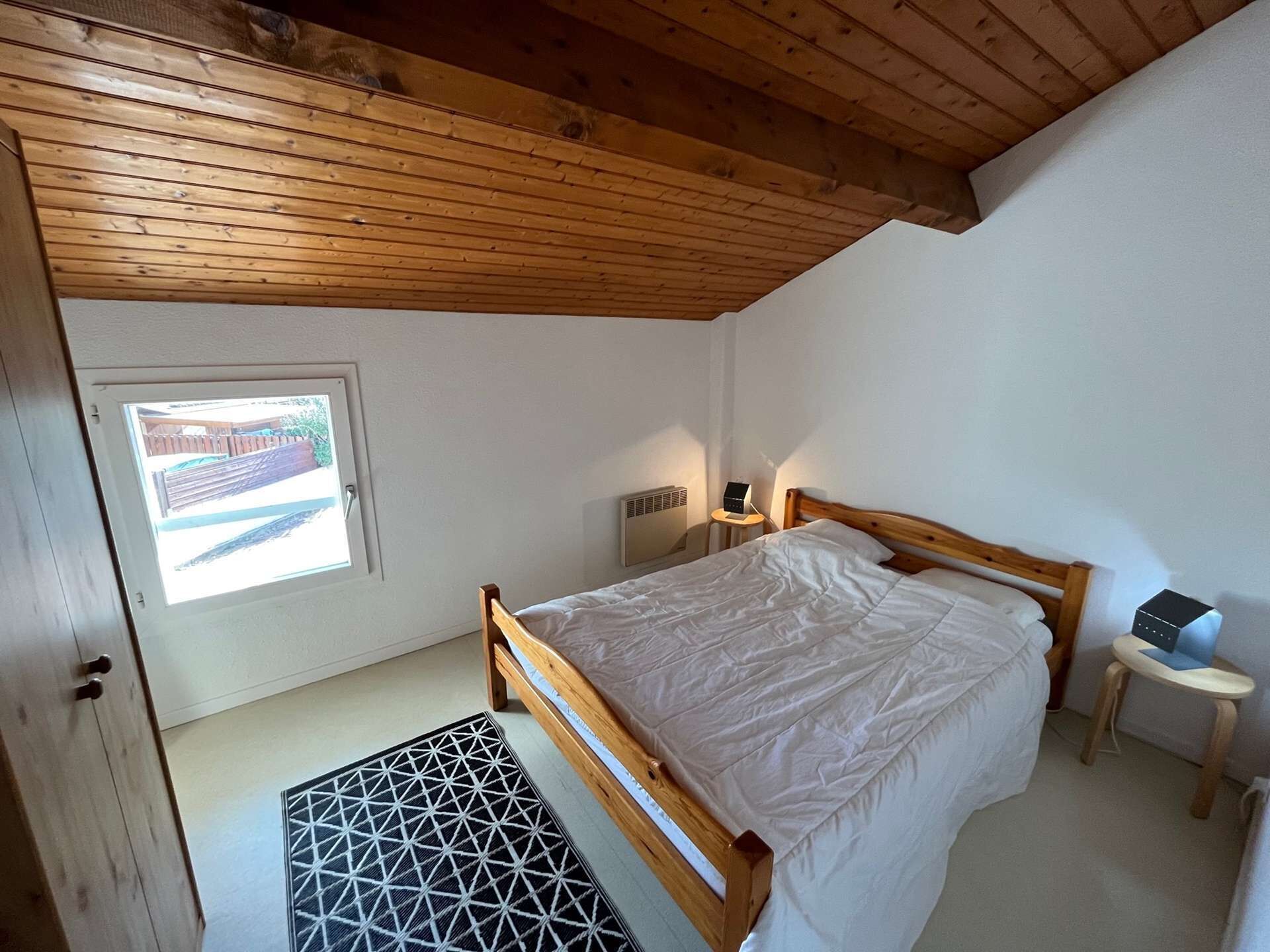 A bedroom with a wooden ceiling, a single bed, a small bedside table with a lamp, a radiator, and a window with a view of a fence.