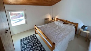 Une chambre avec un plafond en bois, un lit simple, une petite table de chevet avec une lampe, un radiateur et une fenêtre donnant sur une clôture.